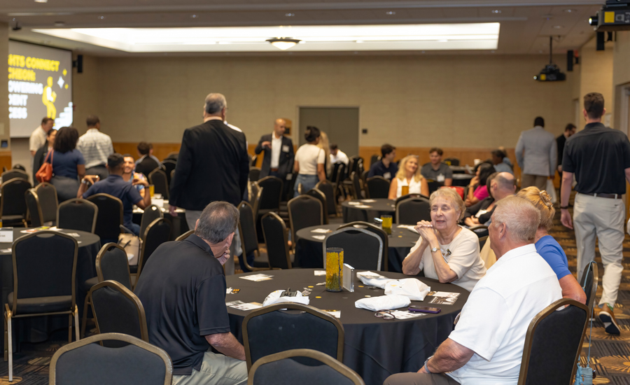 People mingle at a conference reception around round tables in a ballroom setting before a presentation.