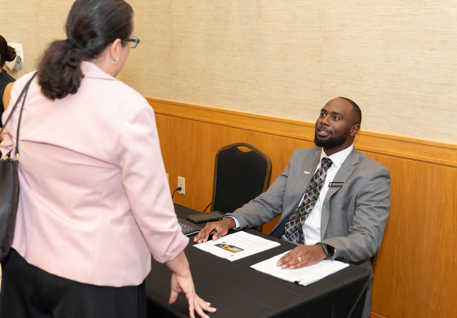 Man in a gray suit sits at a registration desk, greeting a woman in a pink blazer approaching the table.