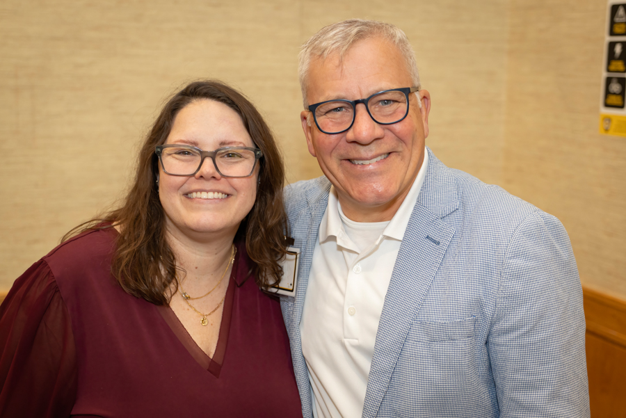 Two smiling adults pose together for a photo at an event, wearing glasses and business casual attire.
