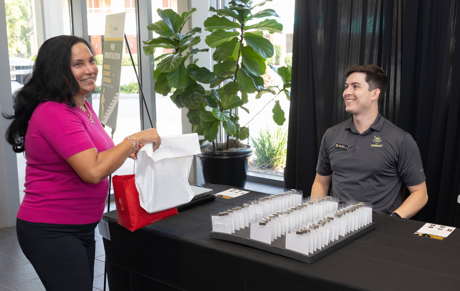 Smiling woman in pink hands a white registration bag to a staff member at a black-draped registration table with name badges on display nearby.