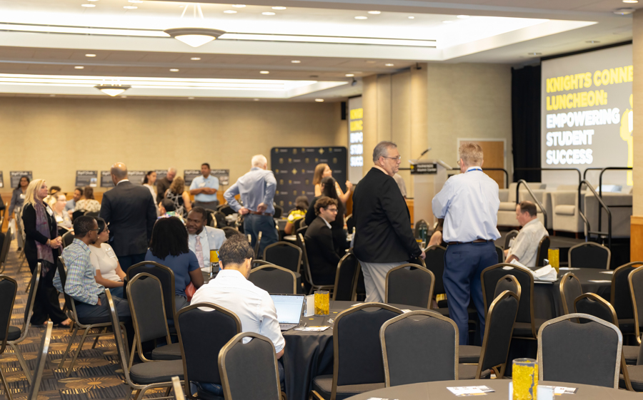 A conference luncheon in a banquet hall with professionals mingling around round tables and a stage with a large screen displaying empowering student‑success messaging in the background.