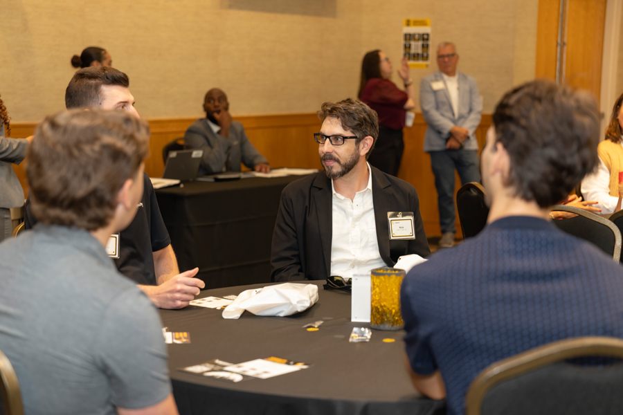 Professionals sit around a round table at a conference, trading ideas and networking with name badges on their chests.