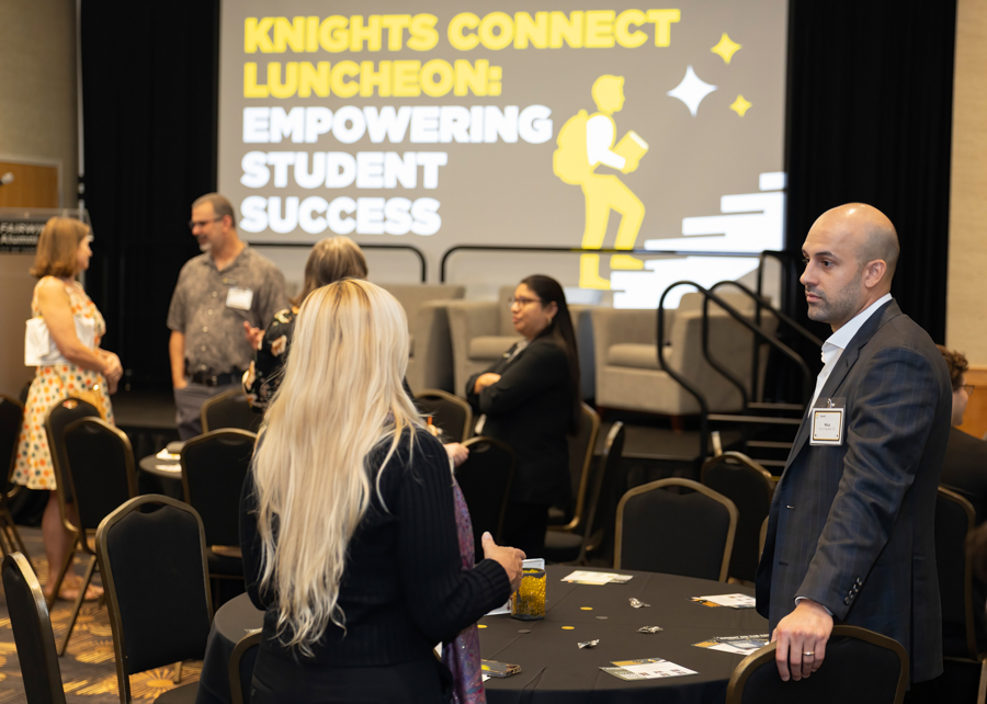 Networking event in a conference room; stage screen reads 'Knights Connect Luncheon: Empowering Student Success' while attendees chat near round tables.
