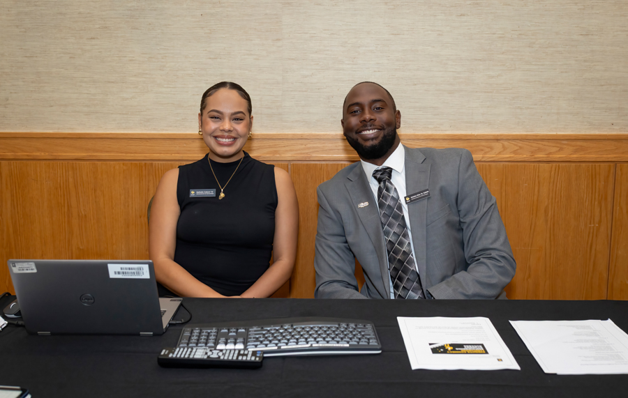 Two smiling staff members sit at a desk with a laptop, keyboard, and papers in front of them on a front desk setup.
