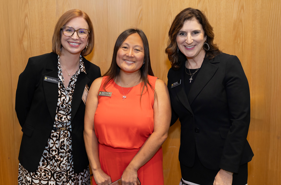 Three smiling professional women posing together in front of a wood-paneled wall at a formal event.