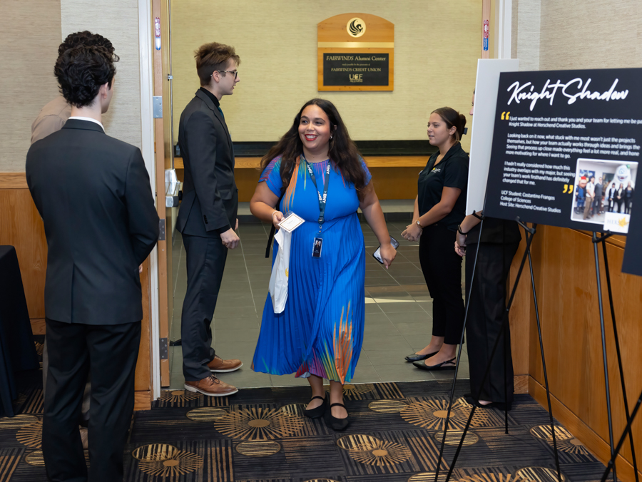 A smiling woman in a blue dress with a lanyard walks through a doorway as two suited men and a woman stand nearby, with a display board on the right.