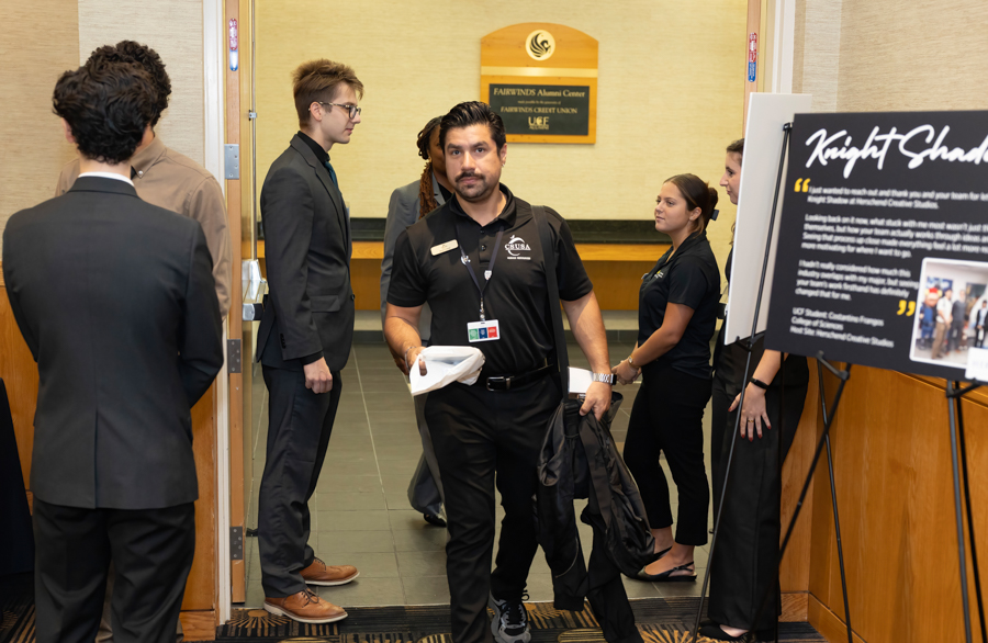 Male staff member in a black polo with a lanyard exits a room carrying a jacket and papers while others stand nearby in a hallway of a campus building.