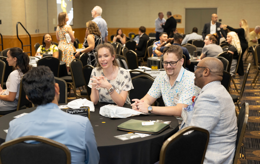 Group of four professionals seated at a round table chatting at a conference event, name badges visible and papers on the table