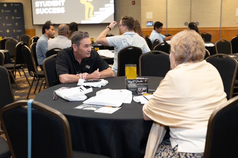 Man in a black polo chats with an elderly woman at a round table during a conference; other attendees sit at tables in the background with a 'Student Success' screen.