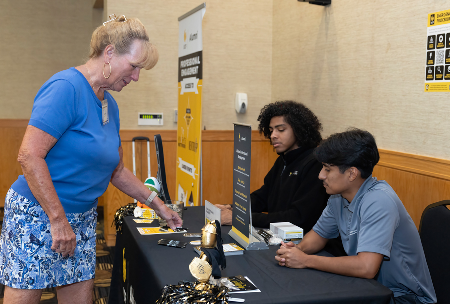Older woman in a blue shirt registers at a black-table booth staffed by two students during an information fair.