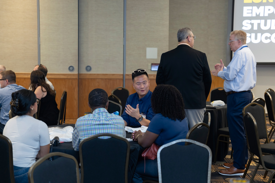 Group of professionals seated around round tables in a conference room, chatting while two men stand near a presentation screen in the background