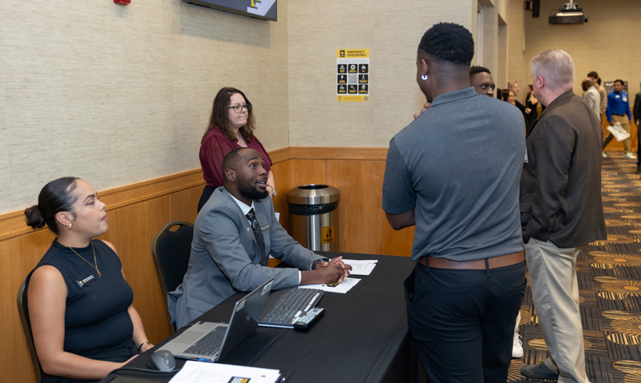 Conference check-in desk: a bearded man in a gray suit sits with two laptops, assisting a standing attendee in a gray shirt as a woman watches nearby.