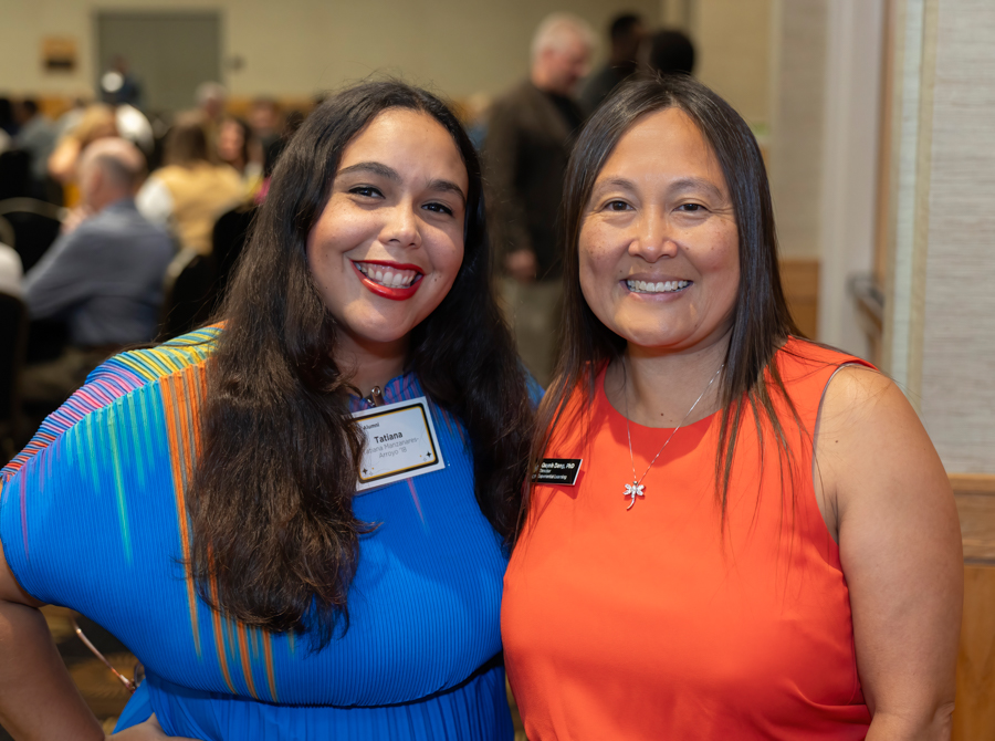 Two smiling women posing at an indoor event; one in a blue dress with a 'Tatiana' name tag, the other in an orange dress with a pendant.