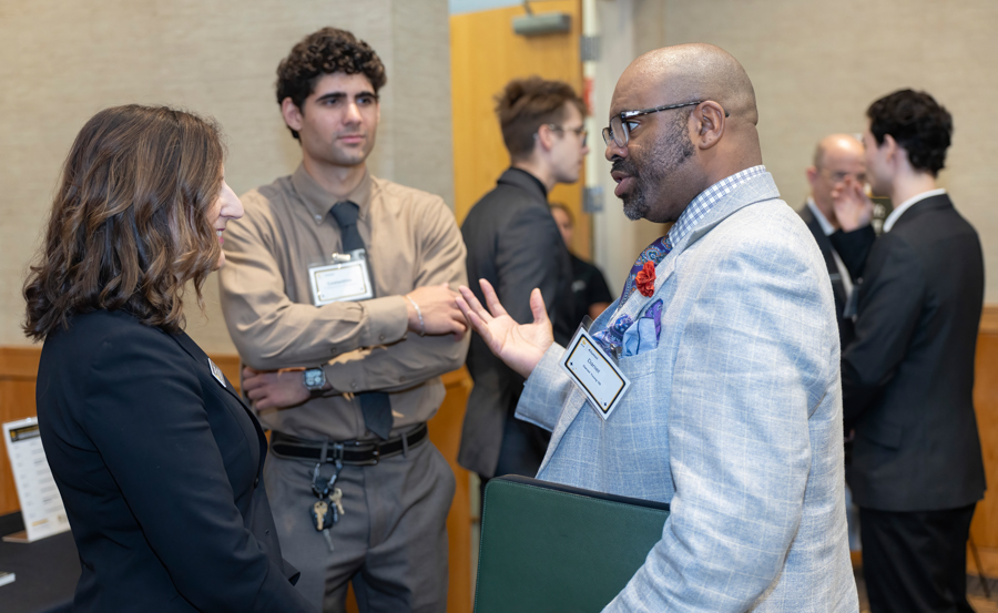 Two professionals in a hallway chat during a networking event, one gesturing with his hand while the other listens.
