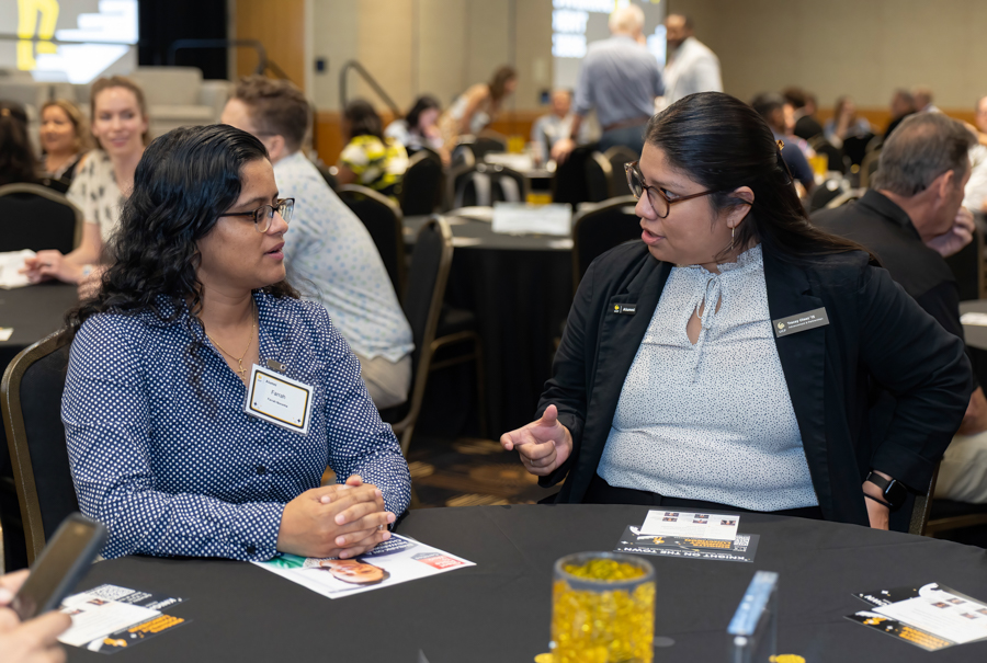 Two women in business attire sit at a round table and discuss at a conference, name badges visible in view.”,