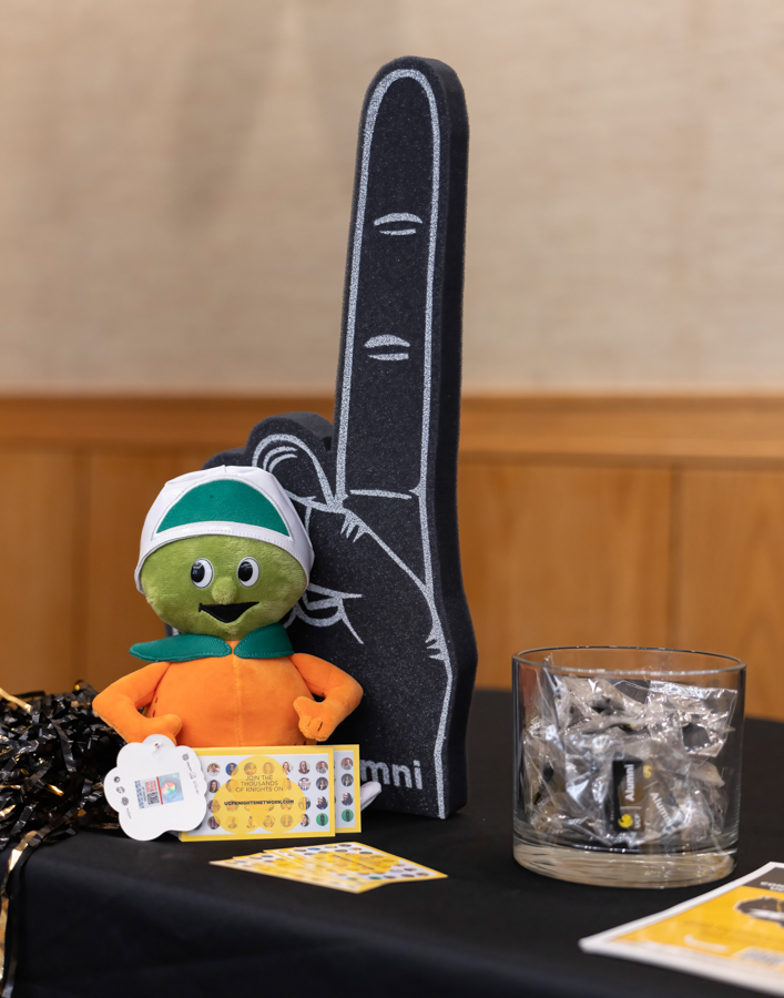 Promotional display with a large foam finger, a green plush mascot in an orange shirt, raffle cards, and a glass bowl of wrapped prizes on a black table.