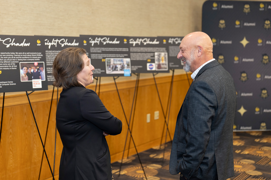 Two professionals in business attire talk in front of exhibition boards at a conference with photos and text on display