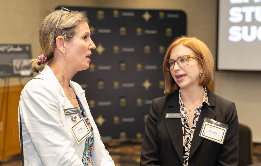 Two professional women in conversation at a conference, wearing name badges in front of a backdrop. (Informative portrait)
