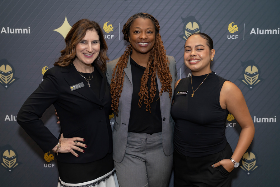 Three women in smart attire posing together at a UCF Alumni backdrop with logos, smiling at the camera.