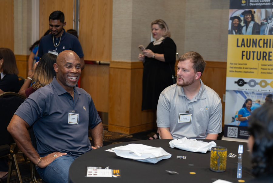 Two men wearing name badges sit at a round table in a conference room, one smiling broadly while the other looks on, with a poster in the background.