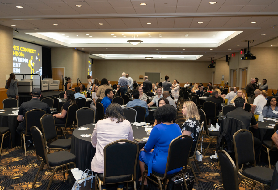 Large conference hall with round tables and attendees networking near a stage with a screen on the left.