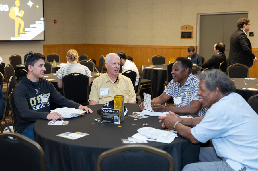 Four men with name badges sit around a round table in a conference room, chatting with papers and drinks on the table.