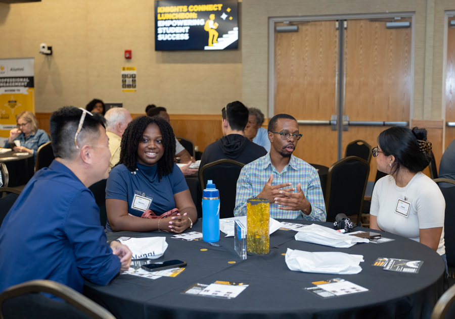 Diverse group of adults seated around a round table at a conference, chatting and taking notes with name badges visible.