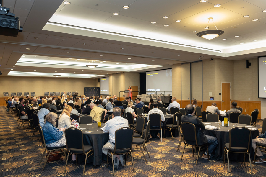 Conference room filled with round tables and attendees watching a speaker at the podium on stage.