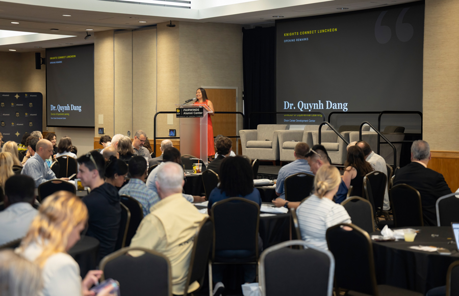 Speaker at a podium on stage addressing attendees at the Knights Connect Luncheon, with a screen displaying 'Dr. Quynh Dang' behind.