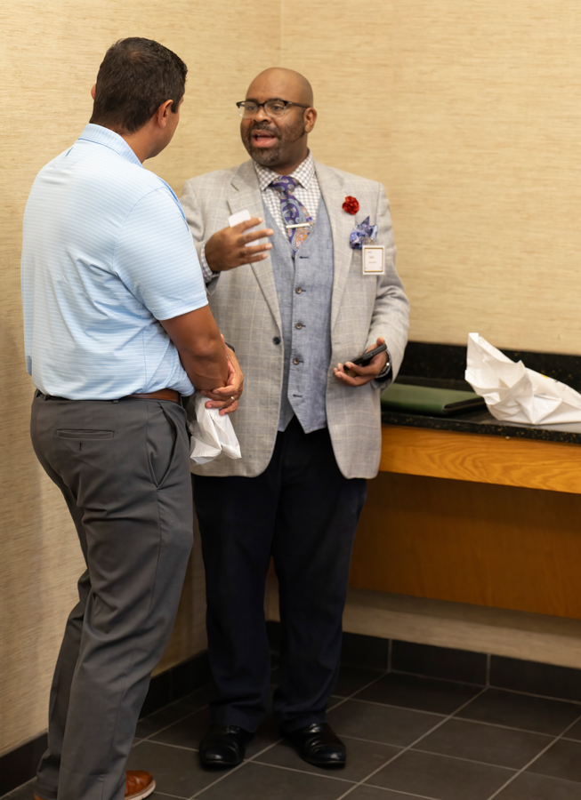 Two men in a hallway conversation: one in a light gray suit with a name badge speaks and gestures, while the other wears a light blue polo and gray pants.