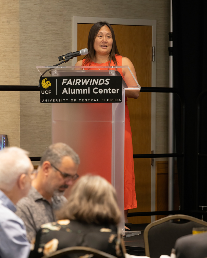 Woman in a red dress speaks at a clear lectern with a microphone at the Fairwinds Alumni Center, UCF signage visible on the podium.