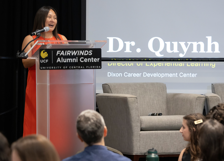 Female speaker in an orange dress at a clear podium with 'FAIRWINDS Alumni Center' branding, addressing an audience as a large screen shows 'Dr. Quynh' in the background.