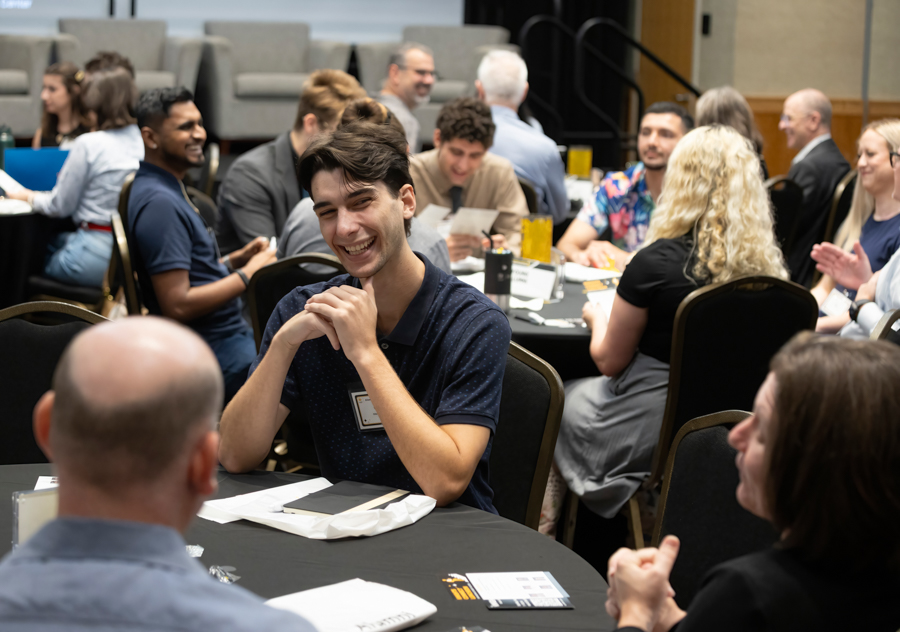 Young man with a name tag sits at a round table during a networking event, smiling and chatting with others around him.