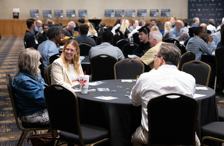 People sit around round tables chatting at a busy conference or networking event in a large room.