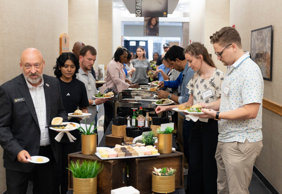 Diverse attendees line up at a buffet in a conference hallway, serving themselves plates from trays with condiments and desserts on display.