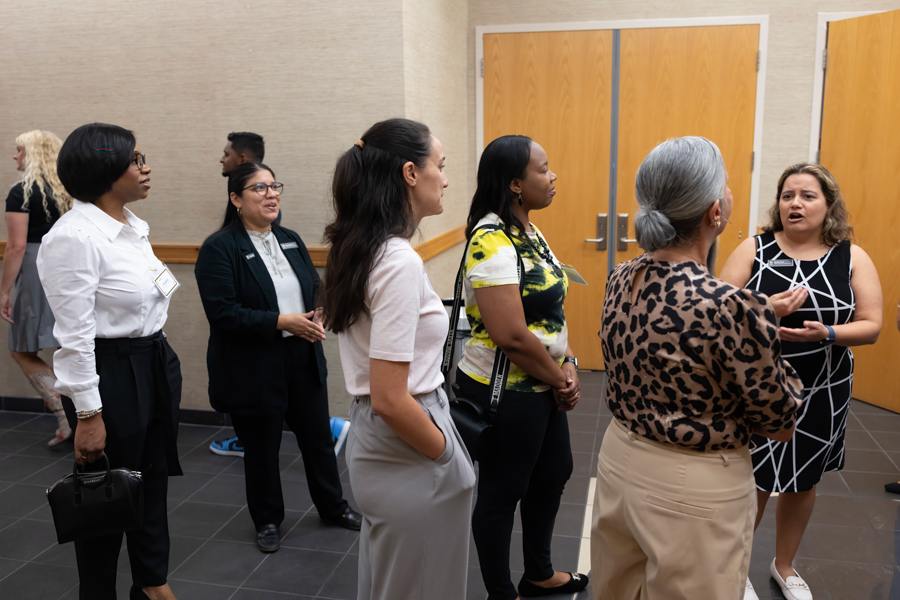 Group of professionals in business casual attire chat in a hallway near wooden doors, some with name badges and lanyards.