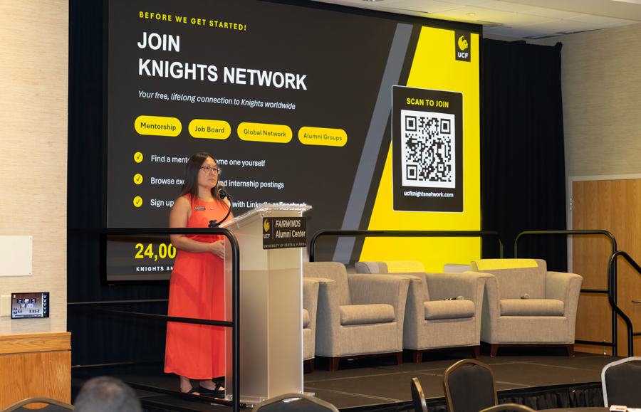 Speaker at a podium on a stage with a large banner that says 'JOIN KNIGHTS NETWORK' and a QR code on the right side of the banner.