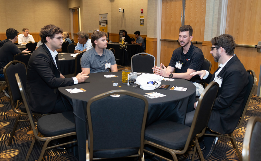 Four men in business attire sit around a round table, talking at a conference or networking event in a hotel meeting room, with name badges visible.