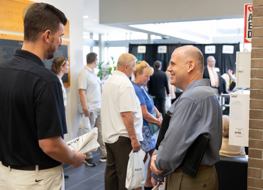 Two men converse at an indoor event; one holds papers as others line up in the background with bags in hand.