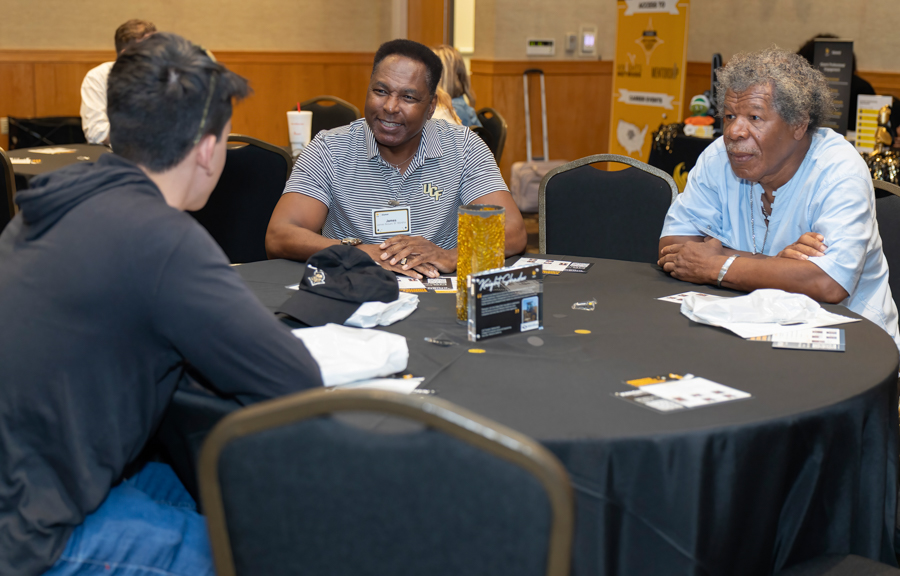 Three people sit around a round table at a conference, chatting and smiling as they network.
