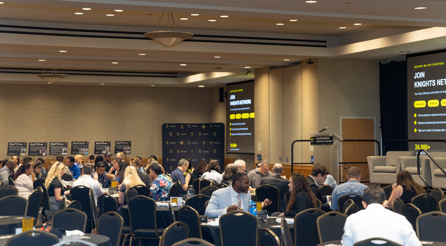Large conference room filled with people seated at round tables, networking during an event; a stage and screens are visible at the front right.