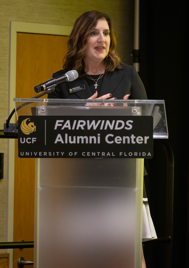 Woman giving a speech at a podium labeled 'Fairwinds Alumni Center' at the University of Central Florida (UCF).
