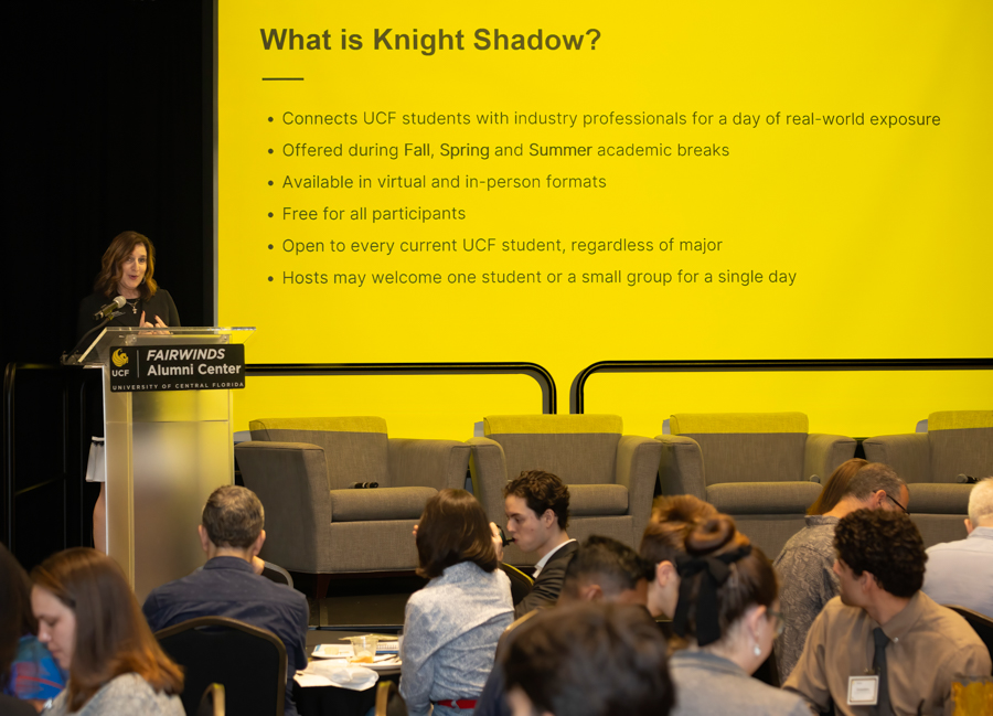 Speaker at a podium presenting to an audience; large yellow slide reads 'What is Knight Shadow?' with bullet points issued.
