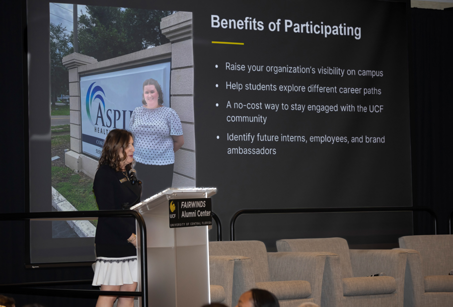 Speaker at a podium presenting a slide titled 'Benefits of Participating' in a conference room.