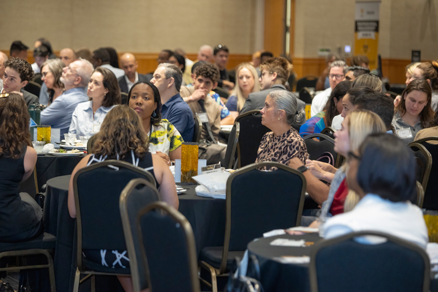 Audience at a formal dinner or conference, seated around round tables listening attentively to a speaker unseen in the photo.