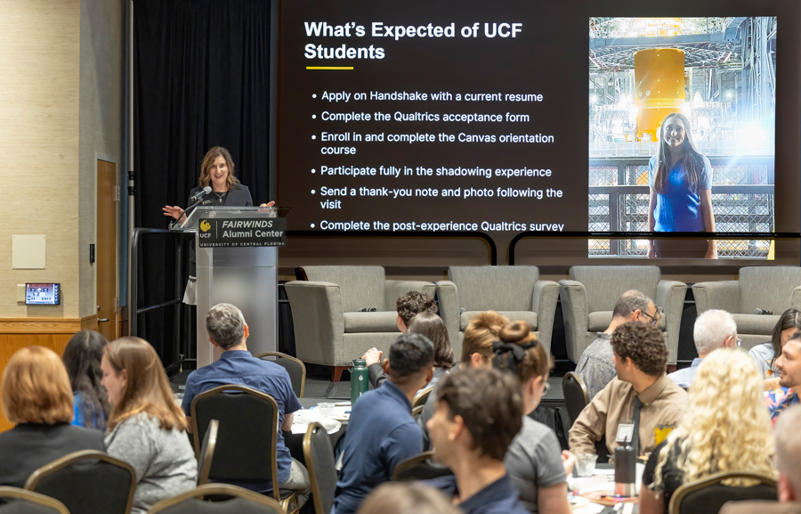 Presenter at a podium on a stage, speaking to a room full of attendees; a large screen behind shows the slide 'What’s Expected of UCF Students' with bullet points and a photo of a person in a blue shirt, and the podium sign reads 'FAIRWINDS Alumni Center'