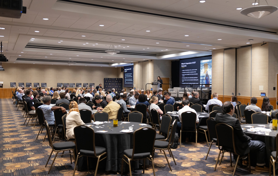 Room filled with conference attendees seated at round tables while a speaker presents at a podium with large presentation screens at the front of a banquet-style ballroom.