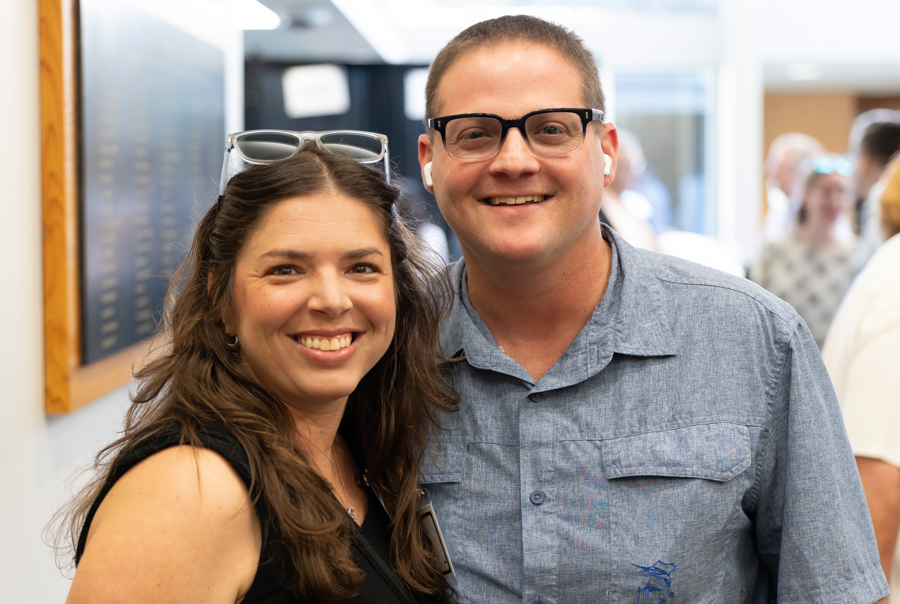 Two smiling adults pose together in a crowded indoor hallway at an event.
