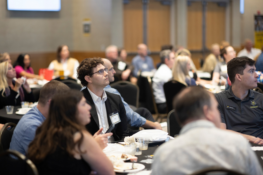Audience seated at round tables in a conference room, paying attention to a presentation.