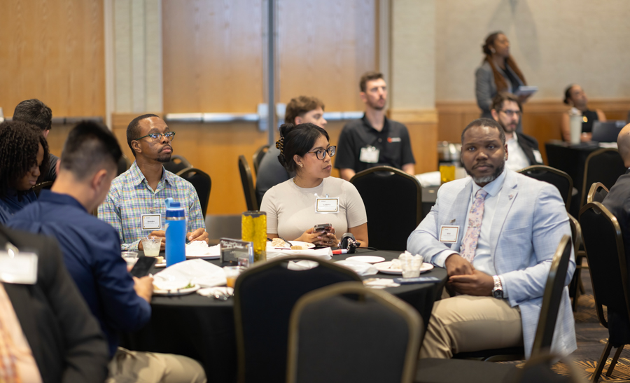 Professionals seated around a round table at a conference, listening and chatting.